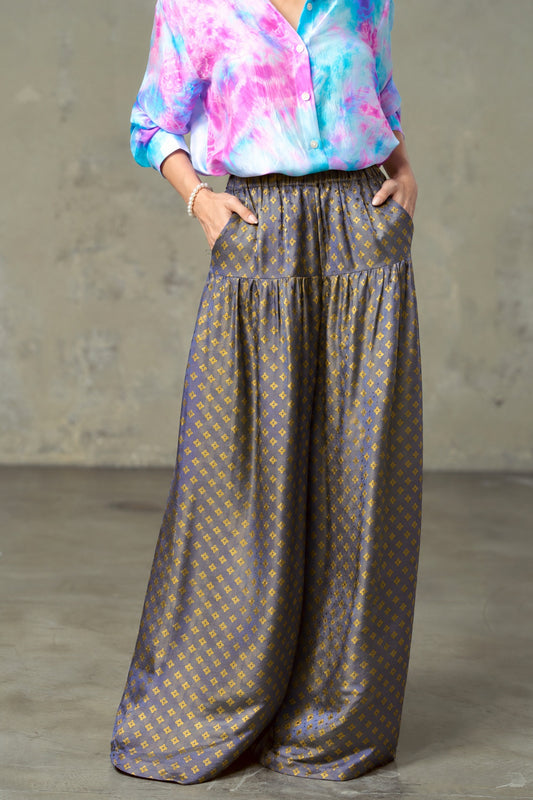 Model wearing Portia Dusk Gold blue and gold silk wide-leg trousers with hands in the side pockets and a tie-dye silk shirt