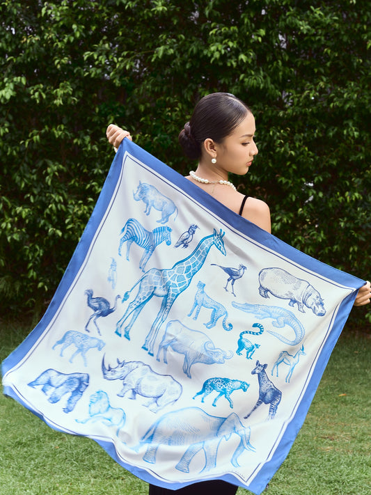 Animal print mulberry silk twill scarf held open, blue engraved giraffe, zebra and elephant on ivory with cornflower border