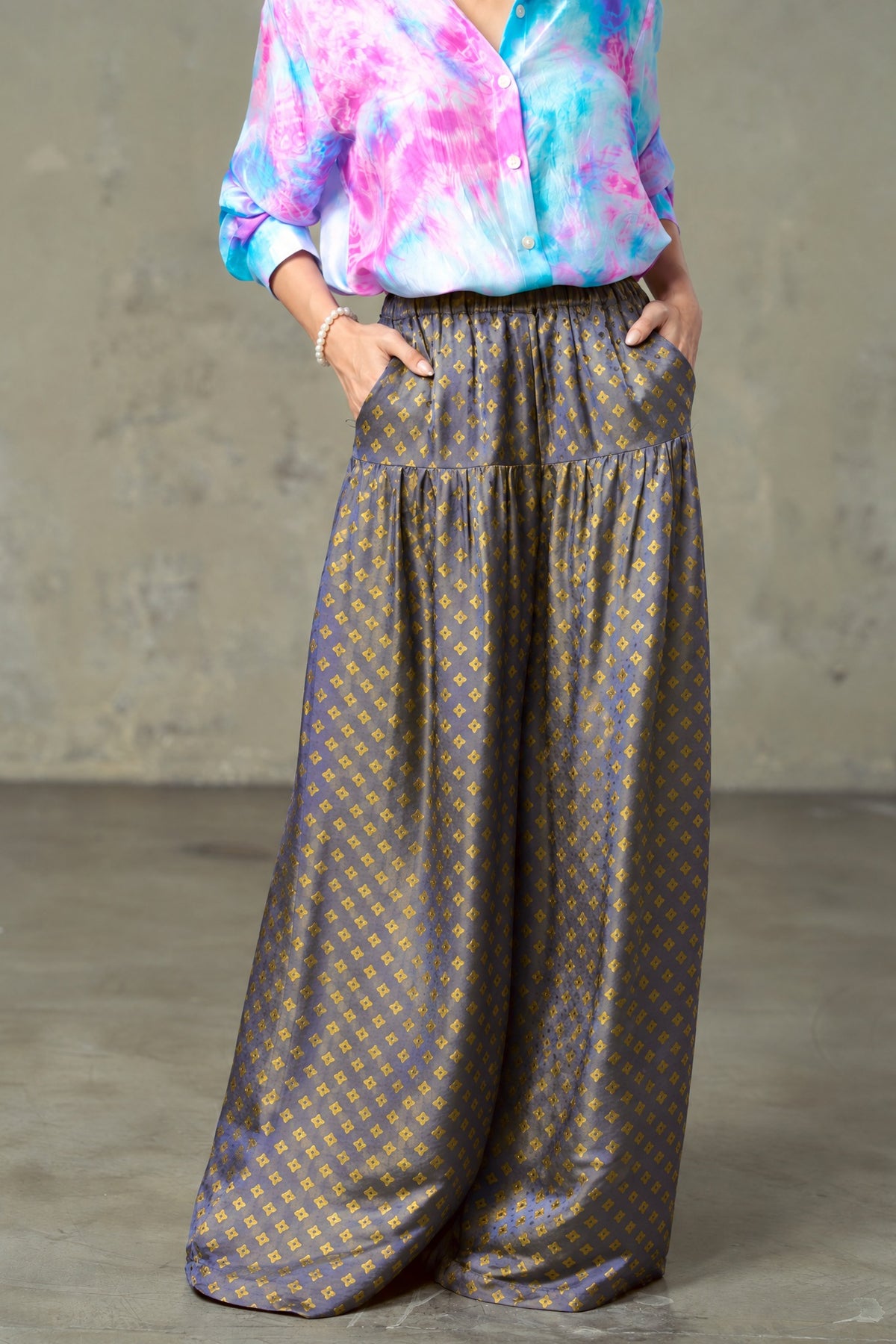 Model wearing Portia Dusk Gold blue and gold silk wide-leg trousers with hands in the side pockets and a tie-dye silk shirt