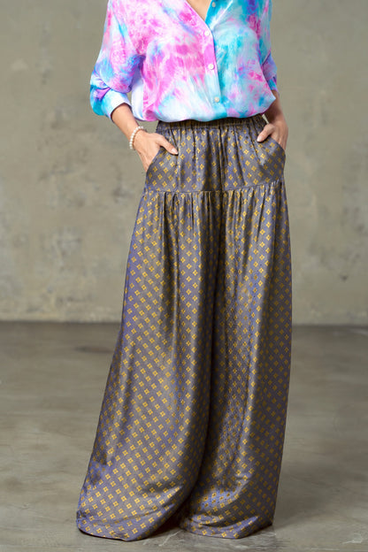 Model wearing Portia Dusk Gold blue and gold silk wide-leg trousers with hands in the side pockets and a tie-dye silk shirt