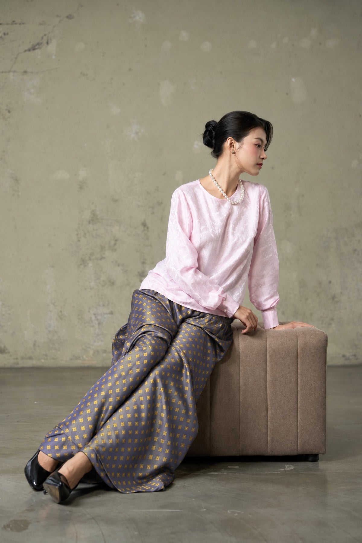 Model seated in Portia Dusk Gold blue-violet and gold silk jacquard wide-leg trousers with a pink silk blouse