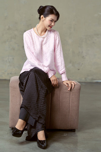 Model seated in blush pink silk jacquard blouse and black jacquard wide-leg trousers